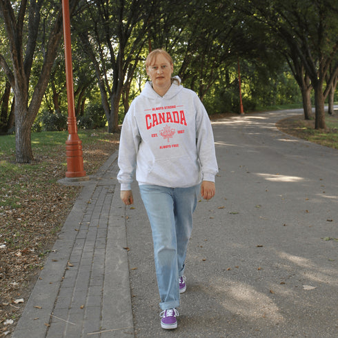 Woman wearing grey Canada Always Strong hoodie with red maple leaf graphic and “Est. 1867” design, styled outdoors in a natural setting