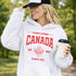 Woman wearing white Canada Always Strong hoodie with red maple leaf graphic and “Est. 1867” design, styled outdoors in a natural setting