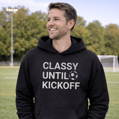 Man wearing “Classy Until Kickoff” hoodie standing on a soccer field during a youth soccer game