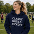 Woman wearing “Classy Until Kickoff” hoodie standing on a soccer field during a youth soccer game