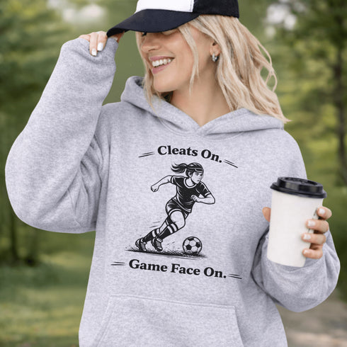 Woman wearing a Cleats On Game Face On soccer hoodie with player graphic, holding coffee outdoors