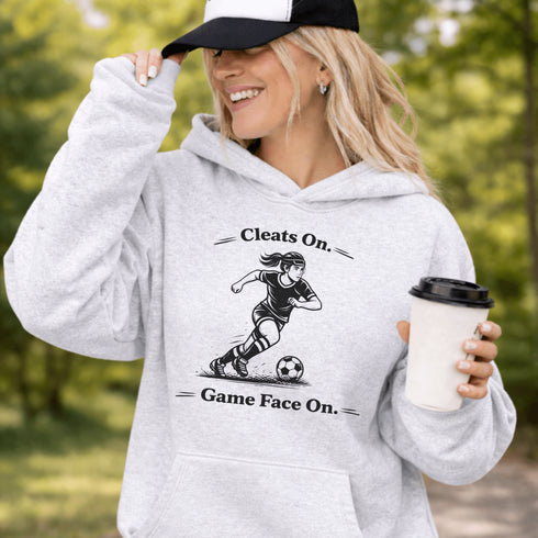 Woman wearing a Cleats On Game Face On soccer hoodie with player graphic, holding coffee outdoors