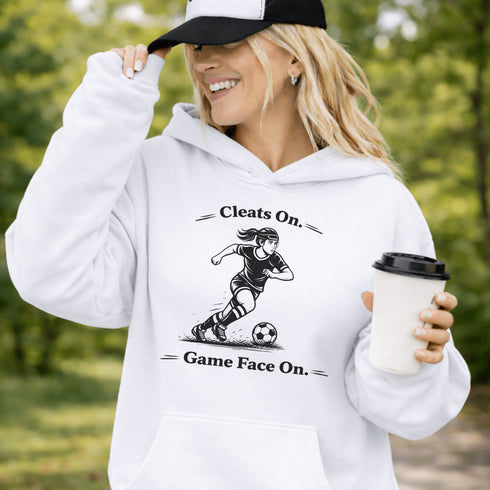 Woman wearing a Cleats On Game Face On soccer hoodie with player graphic, holding coffee outdoors