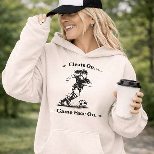 Woman wearing a Cleats On Game Face On soccer hoodie with player graphic, holding coffee outdoors