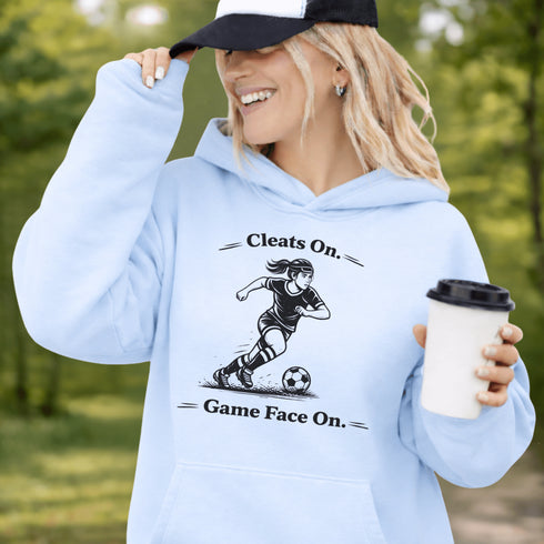 Woman wearing a light blue Cleats On Game Face On soccer hoodie with player graphic, holding coffee outdoors