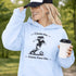 Woman wearing a light blue Cleats On Game Face On soccer hoodie with player graphic, holding coffee outdoors