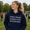 Woman wearing hoodie with "Crazy Proud Always Loud Soccer Mom" text standing on a soccer field during a youth soccer game