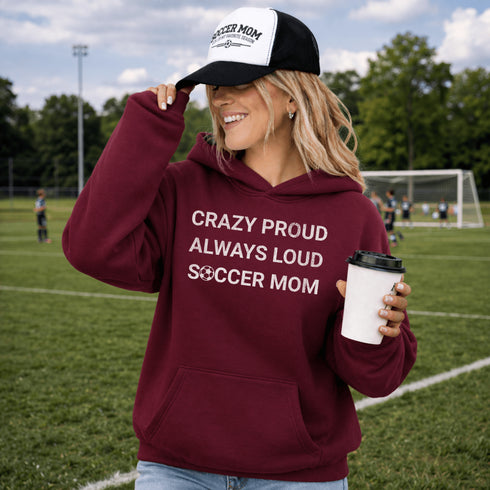 Woman wearing a maroon hoodie with 'Crazy Proud Always Loud Soccer Mom' text, holding a coffee cup on a soccer field.