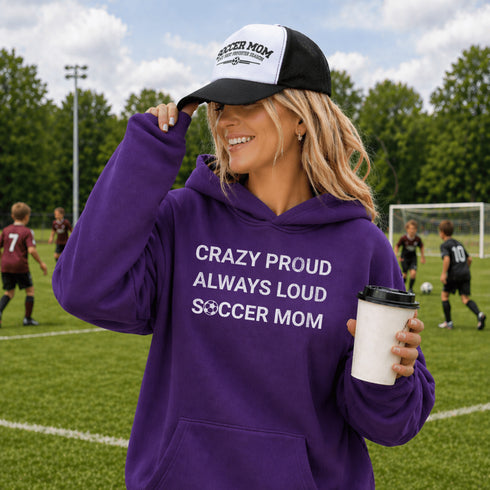 Woman wearing hoodie with "Crazy Proud Always Loud Soccer Mom" text standing on a soccer field during a youth soccer game