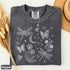 Comfort Colors garment-dyed t-shirt featuring a vintage botanical design with dragonflies, butterflies, bees, wildflowers, dandelions, floating seeds, hearts, stars, and a crescent moon.