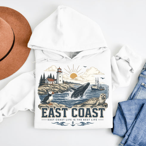 Hoodie with coastal design featuring a lighthouse, whale, and penguin, surrounded by a hat and jeans on a white background.