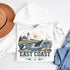 Hoodie with coastal design featuring a lighthouse, whale, and penguin, surrounded by a hat and jeans on a white background.