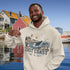 Man wearing a hoodie with a coastal design and text, standing in front of colorful houses by a waterfront.