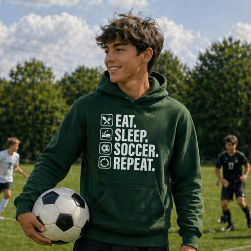 Teen wearing “Eat Sleep Soccer Repeat” hoodie holding a soccer ball on a soccer field during a game, casual soccer apparel