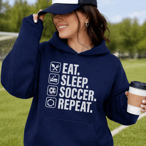 Woman wearing a navy blue “Eat Sleep Soccer Repeat” hoodie holding a coffee on a soccer field during a game, casual soccer apparel