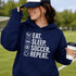 Woman wearing a navy blue “Eat Sleep Soccer Repeat” hoodie holding a coffee on a soccer field during a game, casual soccer apparel