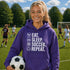 Teen wearing “Eat Sleep Soccer Repeat” hoodie holding a soccer ball on a soccer field during a game, casual soccer apparel