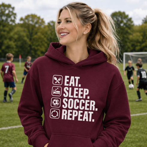 Woman wearing a maroon “Eat Sleep Soccer Repeat” hoodie standing on a soccer field during a game, casual soccer apparel