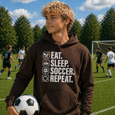 Teen wearing “Eat Sleep Soccer Repeat” hoodie holding a soccer ball on a soccer field during a game, casual soccer apparel