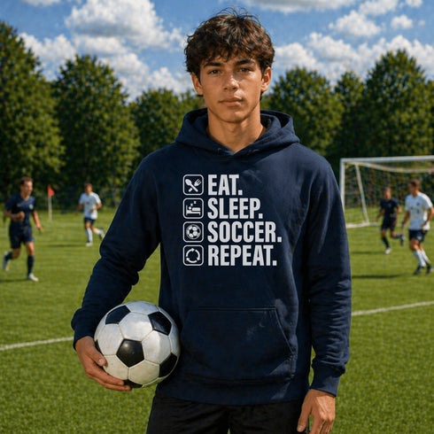 Teen wearing “Eat Sleep Soccer Repeat” hoodie holding a soccer ball on a soccer field during a game, casual soccer apparel
