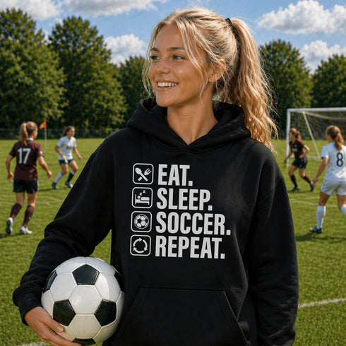Teen wearing “Eat Sleep Soccer Repeat” hoodie holding a soccer ball on a soccer field during a game, casual soccer apparel