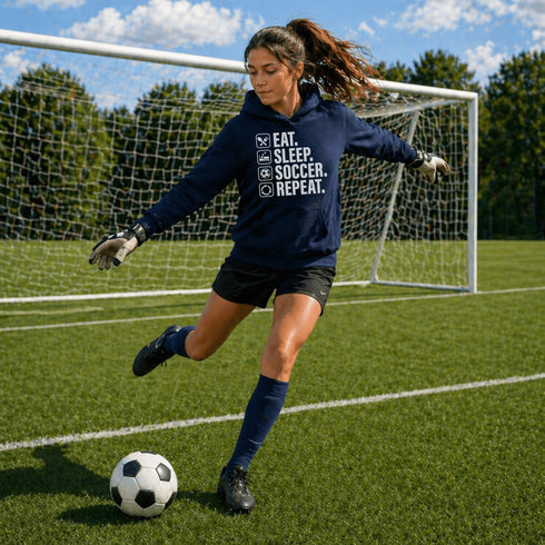 Female teen goalie wearing a navy blue “Eat Sleep Soccer Repeat” hoodie kicking a soccer ball on a soccer field during a game, casual soccer apparel