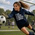 Female teen goalie wearing a navy blue “Eat Sleep Soccer Repeat” hoodie saving a soccer ball on a soccer field during a game, casual soccer apparel