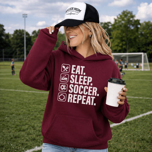 Woman wearing a maroon “Eat Sleep Soccer Repeat” hoodie holding a coffee on a soccer field during a game, casual soccer apparel