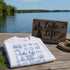 White t-shirt with 'Lake Life' design on a wooden dock by a lake.