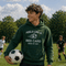 Person wearing a green hoodie with a soccer-themed quote on a soccer field