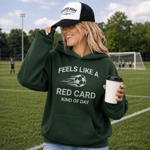 Woman wearing a green hoodie with text and a soccer ball graphic, holding a coffee cup on a soccer field.