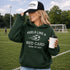 Woman wearing a green hoodie with text and a soccer ball graphic, holding a coffee cup on a soccer field.