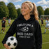 Woman on a soccer field wearing a black hoodie with a humorous soccer-themed quote, holding a soccer ball.