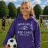 Woman in purple hoodie holding a soccer ball on a soccer field with other players in the background.