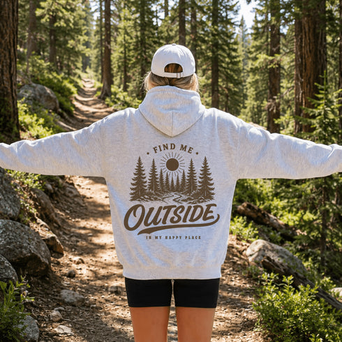 A woman standing on a forest trail with her arms outstretched, wearing a light heather gray hoodie with a brown nature graphic print that reads 'FIND ME OUTSIDE IN MY HAPPY PLACE' and a white baseball cap.
