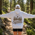 A woman standing on a forest trail with her arms outstretched, wearing a light heather gray hoodie with a brown nature graphic print that reads 'FIND ME OUTSIDE IN MY HAPPY PLACE' and a white baseball cap.
