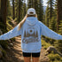 A woman standing on a forest trail with her arms outstretched, wearing a light blue hoodie with a large brown back nature graphic that reads “FIND ME OUTSIDE IN MY HAPPY PLACE,” along with a white cap and black shorts.