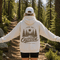 A woman stands on a forest trail with her arms outstretched, wearing a white hoodie from the back. The hoodie features a large brown graphic of pine trees, a sun, and the words 'FIND ME OUTSIDE IN MY HAPPY PLACE.