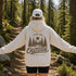 A woman stands on a forest trail with her arms outstretched, wearing a white hoodie from the back. The hoodie features a large brown graphic of pine trees, a sun, and the words 'FIND ME OUTSIDE IN MY HAPPY PLACE.