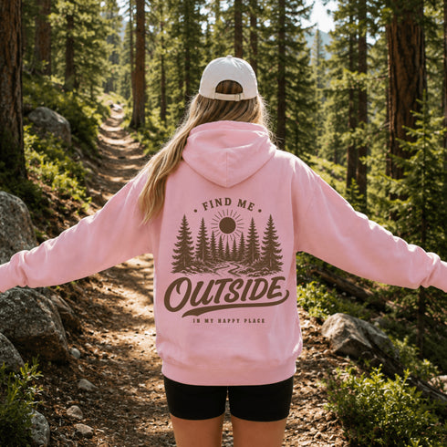 A woman standing on a forest trail seen from behind, wearing a light pink hoodie with a large brown nature graphic on the back that reads “FIND ME OUTSIDE IN MY HAPPY PLACE,” along with a white cap and black shorts.
