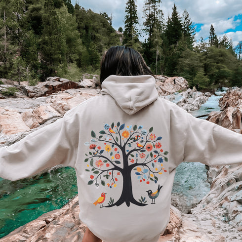 This hoodie features a delightful folk art design of vibrant birds perched in a tree adorned with colorful leaves and blooming flowers, creating a scene that radiates joy and creativity.