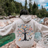 This hoodie features a delightful folk art design of vibrant birds perched in a tree adorned with colorful leaves and blooming flowers, creating a scene that radiates joy and creativity.