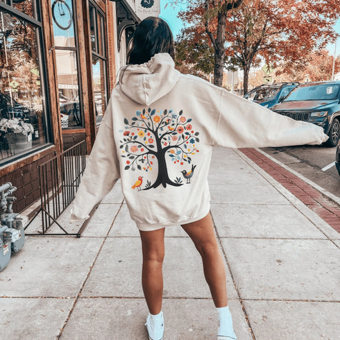 This hoodie features a delightful folk art design of vibrant birds perched in a tree adorned with colorful leaves and blooming flowers, creating a scene that radiates joy and creativity.