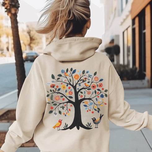 This hoodie features a delightful folk art design of vibrant birds perched in a tree adorned with colorful leaves and blooming flowers, creating a scene that radiates joy and creativity.