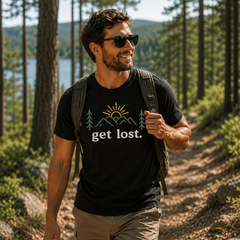 Man hiking on a forest trail overlooking a lake while wearing a black Get Lost t-shirt featuring a mountain and sunrise outdoor design.