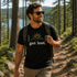 Man hiking on a forest trail overlooking a lake while wearing a black Get Lost t-shirt featuring a mountain and sunrise outdoor design.
