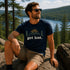 Man sitting on a rock with a scenic view of a lake and forest, wearing a navy t-shirt with 'get lost.' text.