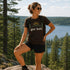 Woman wearing a 'get lost' t-shirt in a scenic outdoor setting with trees and water.