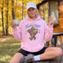 A woman sitting outdoors on a wooden deck in front of a cabin, wearing a light pink hoodie with a cartoon Bigfoot graphic and the text 'GONE TILL Further Notice.' She holds a white mug in one hand and makes a peace sign with the other.