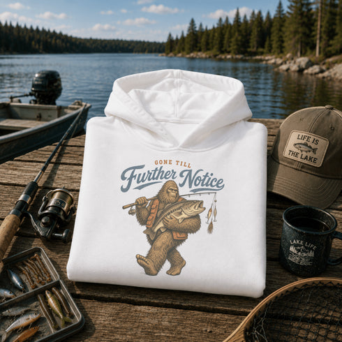 A white pullover hoodie is folded on a wooden dock with a lake and forest in the background. The hoodie has a graphic print of Bigfoot holding a fish with the text 'GONE TILL Further Notice.